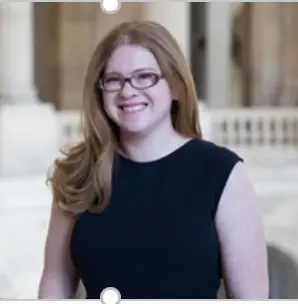 A woman with long, light brown hair and glasses is smiling at the camera. She is wearing a sleeveless black dress and standing in a bright, possibly governmental building with white columns and railings in the background, highlighting her involvement in CMMC initiatives to combat cyberthreats within the DOD.