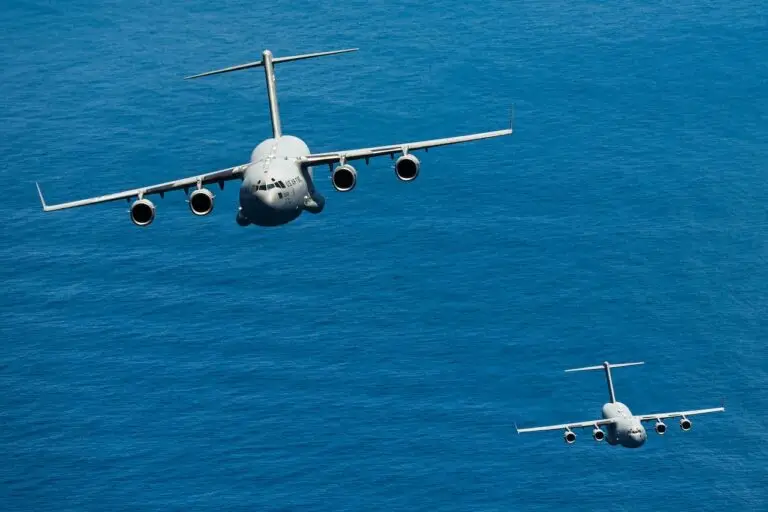 Two military cargo planes, laden with cybersecurity equipment to counter cyberthreats, fly in formation over a vast, deep blue ocean. The larger lead aircraft is closest to the camera, while the smaller follows at a lower altitude in the background. Both planes have visible USAF markings.