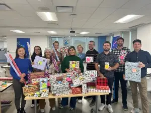 A group of ten people are standing behind a table covered with wrapped gifts and rolls of wrapping paper in an office with fluorescent lighting and large windows. Smiling and holding additional wrapped presents, they look like they're discussing something important—perhaps a new VPN for improving cybersecurity.