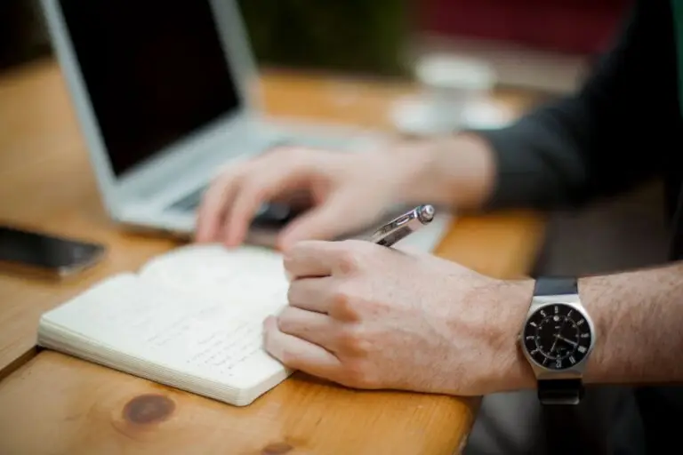 A person with a wristwatch is writing in a notebook with a pen while using a laptop on a wooden table. A smartphone and a saucer with a teacup are also on the table, possibly addressing cyberthreats in their work. The scene suggests a casual work or study environment.