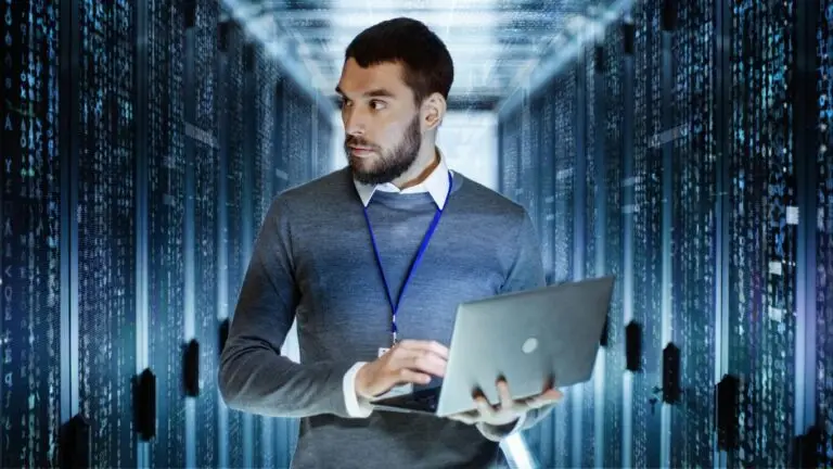 A man wearing a grey sweater and white shirt, holding and working on a laptop, stands in the corridor of a server room. He's looking to his side amidst rows of servers and cables, illuminated by cool-toned lighting. The setting emphasizes the critical importance of cybersecurity in today's CMMC-compliant environments.