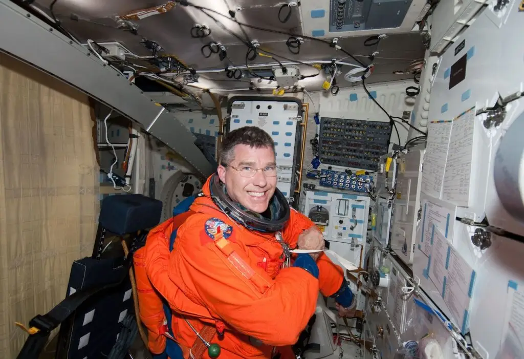 An astronaut in an orange space suit smiles at the camera while seated inside a space shuttle cockpit. The control panel is filled with switches, buttons, and screens. Amidst the wires and equipment creating a busy but organized environment, cybersecurity measures ensure that every system is protected against cyberthreats.