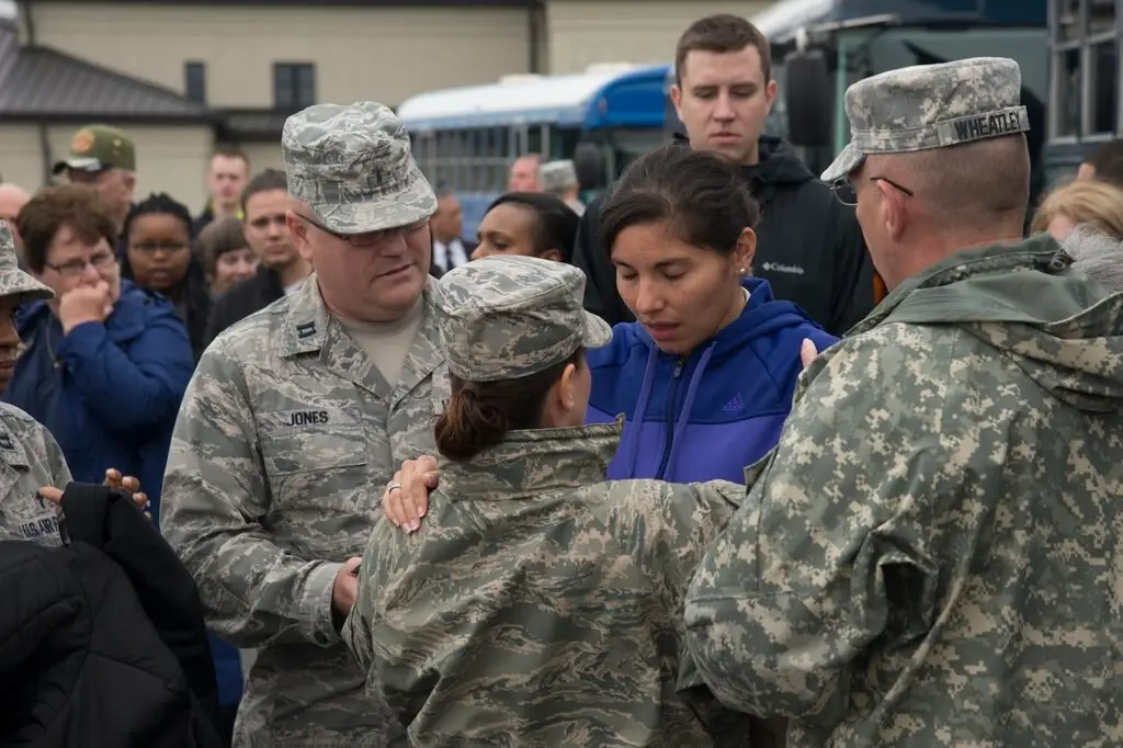 A woman in a blue and purple jacket is surrounded by uniformed military personnel and a crowd. One officer, wearing a camouflage uniform, touches her arm while others look on. They appear to be outdoors near a few buses, possibly discussing a cybersecurity threat with DOD officials.