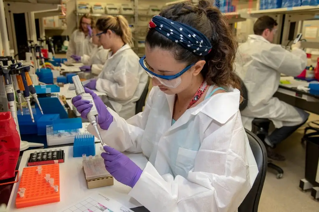 A group of scientists in a laboratory are conducting experiments. The woman in the foreground is wearing a white lab coat, safety goggles, and purple gloves. She is using a pipette to transfer liquid samples into a tray, mindful of potential cybersecurity threats. The lab is equipped with various scientific instruments.