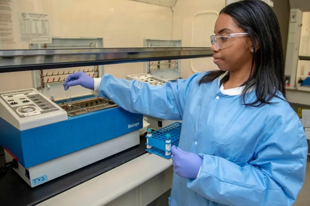 A scientist wearing a blue lab coat, safety glasses, and purple gloves is using laboratory equipment. She is alert to potential threats as she inserts a small vial into a machine with multiple slots, surrounded by various lab instruments and a rack holding test tubes.