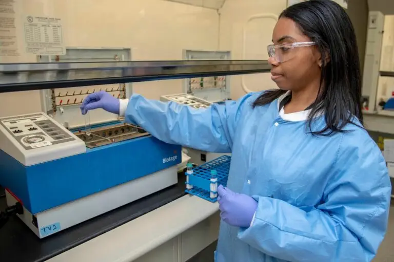 A scientist wearing a blue lab coat, safety glasses, and purple gloves is using laboratory equipment. She is alert to potential threats as she inserts a small vial into a machine with multiple slots, surrounded by various lab instruments and a rack holding test tubes.