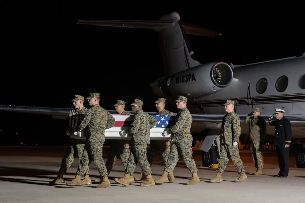 A group of military personnel in camouflage uniforms solemnly carry a casket draped with the American flag from an aircraft on a tarmac at night. A naval officer salutes in the background. The scene, illuminated by artificial lighting, evokes the vigilance required to guard against cyberthreats.