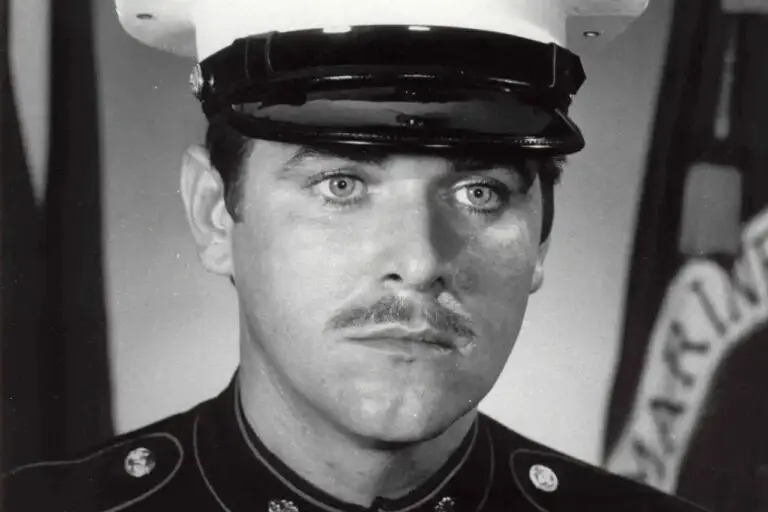 A black and white portrait of a man in military dress uniform and a white peaked cap. The man, with a mustache, looks slightly to his left with a serious expression. The background features a flag with partially visible text, hinting at his role defending against looming cyberthreats for the DOD.