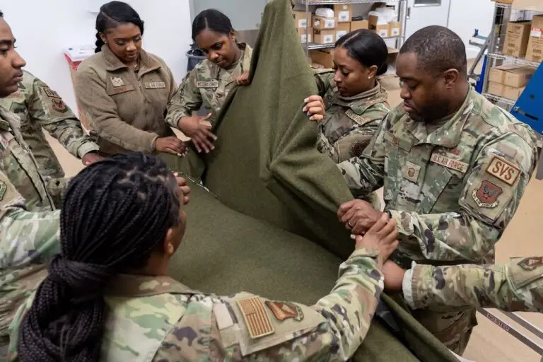 A group of individuals dressed in military uniforms work together to fold a large green fabric. They are standing in a room with shelves containing boxes and other supplies, appearing focused and coordinated as if preparing for a mission against an imminent cyberthreat.