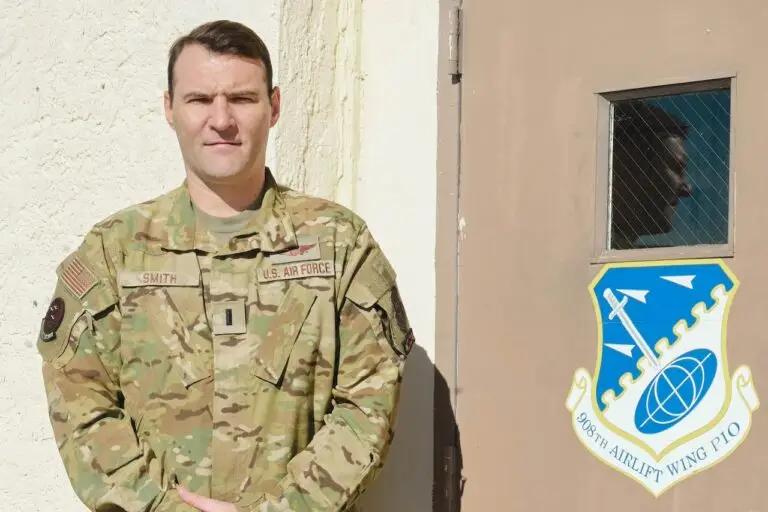 A member of the U.S. Air Force, wearing a camouflage uniform, stands near a building with a sign reading "386th Airlift Wing PIO" and a logo featuring a plane and globe. The individual has a neutral expression while shadows cast by the sun accentuate the wall, highlighting their role in cybersecurity defense for the DOD.