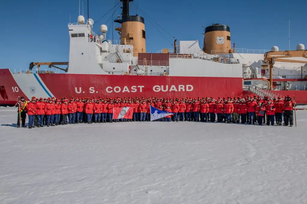 A large group of U.S. Coast Guard personnel in red jackets stands in front of a Coast Guard icebreaker ship on a snowy surface. Some individuals in the front row are holding various flags, including one white flag and another with blue and red elements, demonstrating unity against any potential threat.