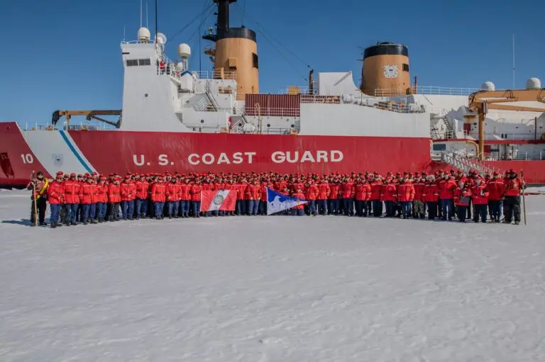 A large group of U.S. Coast Guard personnel in red jackets stands in front of a Coast Guard icebreaker ship on a snowy surface. Some individuals in the front row are holding various flags, including one white flag and another with blue and red elements, demonstrating unity against any potential threat.