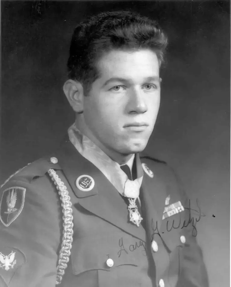 Black and white portrait of a young man in military uniform, adorned with various insignia, medals, and an award around his neck. He has short, neatly styled hair and a serious expression. An autograph is scribbled on the bottom right of the photo, highlighting his role in addressing cyberthreats for the DOD.