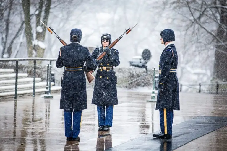 Three uniformed soldiers perform a guard change ceremony in the snow. Two soldiers face each other with rifles at shoulder arms, while a third stands to the side observing. Snowfall and trees are visible in the background, creating a solemn winter scene under strict DOD protocol.