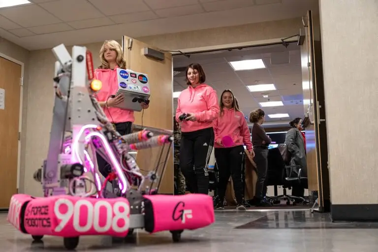 Three people wearing matching pink hoodies are operating a large robotic vehicle with the number 9008 displayed on it. The robot, illuminated by pink LED lights, moves through a hallway. In the background, more people are visible, emphasizing the threat of potential data breaches without proper CMMC protocols in place.