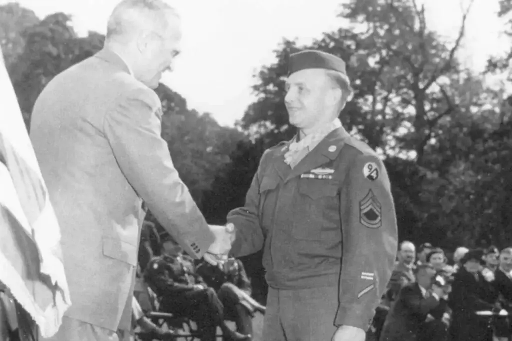 A black-and-white photograph shows a military officer in uniform shaking hands with a man in a suit at a formal outdoor ceremony. Seated onlookers watch, while trees provide a serene backdrop. This event underscores the importance of cybersecurity, highlighting initiatives like CMMC to protect our digital frontiers.