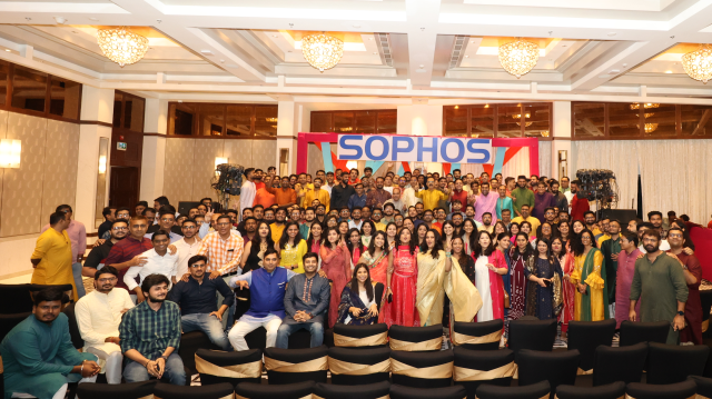 A large group of people, dressed in colorful traditional attire, pose for a group photo in a well-lit hall with chandeliers. The backdrop features the text "SOPHOS" in bold. Everyone appears cheerful and is facing the camera, symbolizing unity against cyber threats.