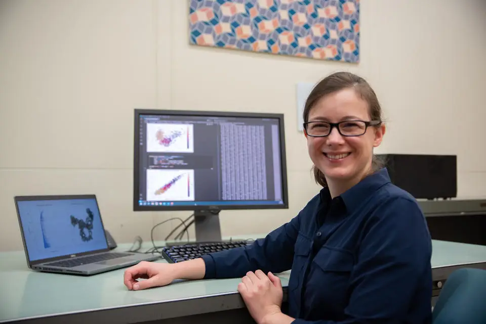 A woman wearing glasses and a dark shirt sits at a desk with a laptop and a monitor displaying data visualizations related to cybersecurity. She is smiling and looking at the camera, with a geometric art piece visible on the wall behind her.