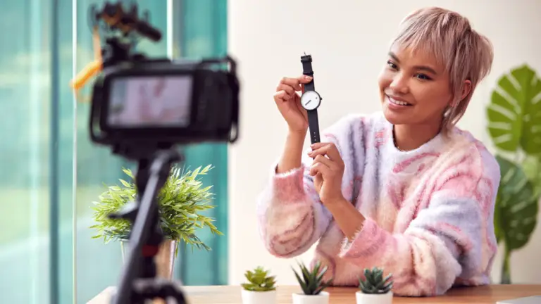 A person with short hair and a colorful sweater showcasing a watch to a camera on a tripod. The background includes large windows, lush plants, and ample natural light, providing an inviting setting that feels secure without any threat to privacy—much like using a reliable VPN.