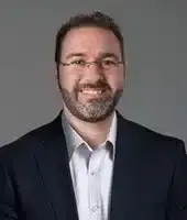 A man with short dark hair, glasses, and a beard smiles at the camera. He is wearing a dark suit jacket over a light-colored dress shirt, perhaps discussing the importance of using a VPN to protect against cyberthreats. The background is a plain, neutral gray.