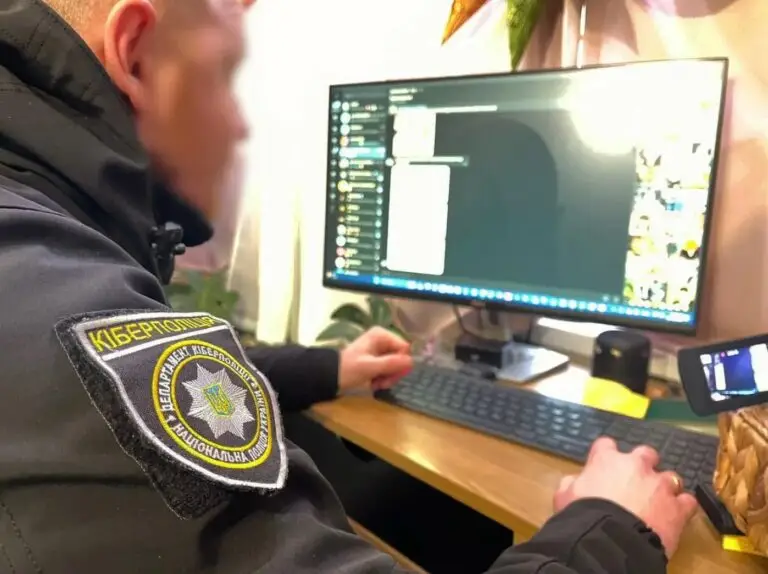 A person in a uniform with a patch reading "Кіберполіція" works on a computer. The monitor displays a messaging interface, and there's a smartphone mounted on a stand next to it. Amidst office items and plants, the scene subtly underscores the importance of cybersecurity against potential threats.