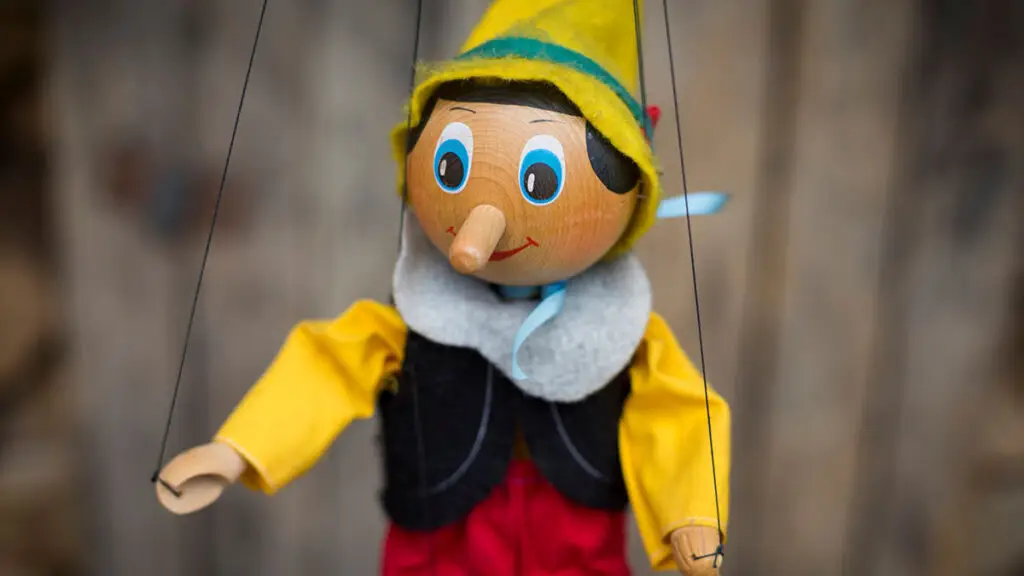 A marionette puppet resembling Pinocchio with a yellow hat, blue bow tie, red trousers, and a blue vest over a yellow shirt is shown. The puppet is suspended by strings and has a cheerful expression. The background, which looks like slightly blurred wood, evokes no hint of threat in its charming simplicity.