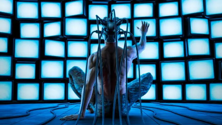 A person with a bare back and camouflage pants sits on the floor facing a wall of glowing screens. They are connected to the screens with cables attached to a headgear resembling a neural interface, possibly fending off a cyberthreat. The blue light from the screens illuminates the scene.