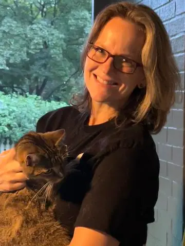 A woman with glasses and shoulder-length hair smiles while holding a tabby cat in her arms. They are standing near a window, and greenery is visible outside. She is wearing a black shirt, perhaps unwinding after completing her work on the DOD's latest threat assessment. The scene appears to be indoors.