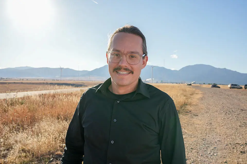 A man with glasses, a mustache, and long hair pulled back is smiling while standing outside on a sunny day. He is wearing a black button-up shirt and standing on a gravel path with mountains, grasslands, wind turbines in the background. His interest in cybersecurity often leads him to consider cyberthreat scenarios while enjoying nature.