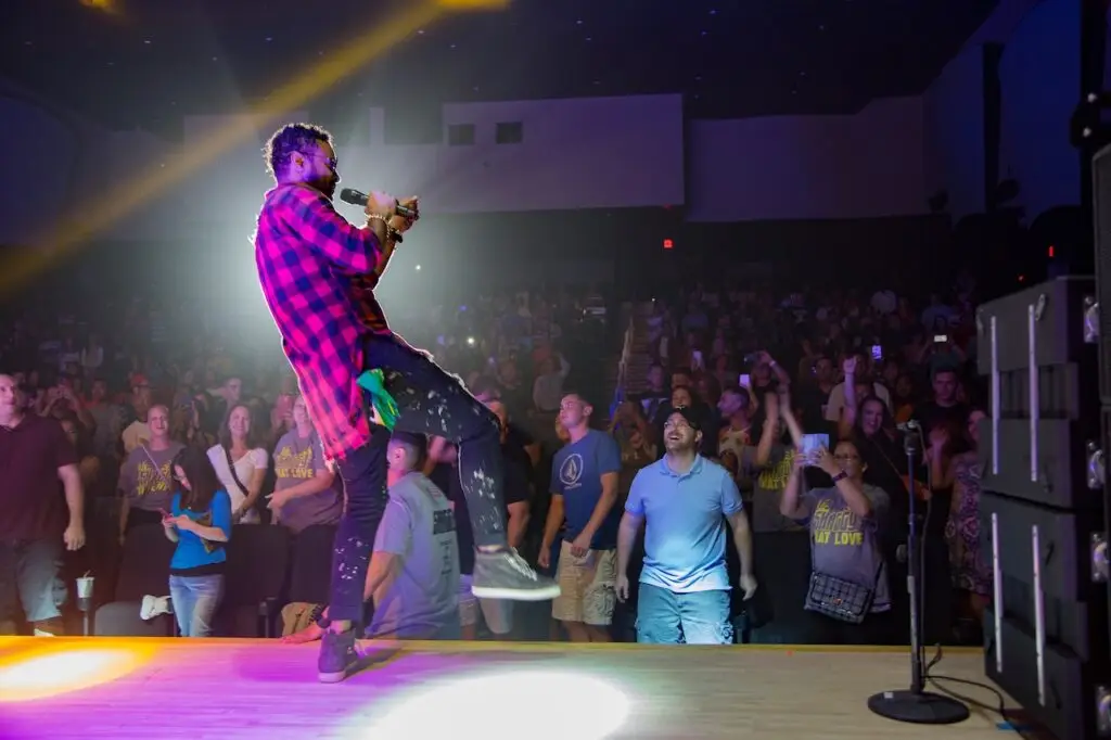 A performer in a red checkered shirt plays an instrument on stage under bright lights, facing a cheering audience. The crowd is standing, clapping, and taking photos, with many people appearing excited. Despite the festive atmosphere, the event had heightened security due to a cyberthreat.