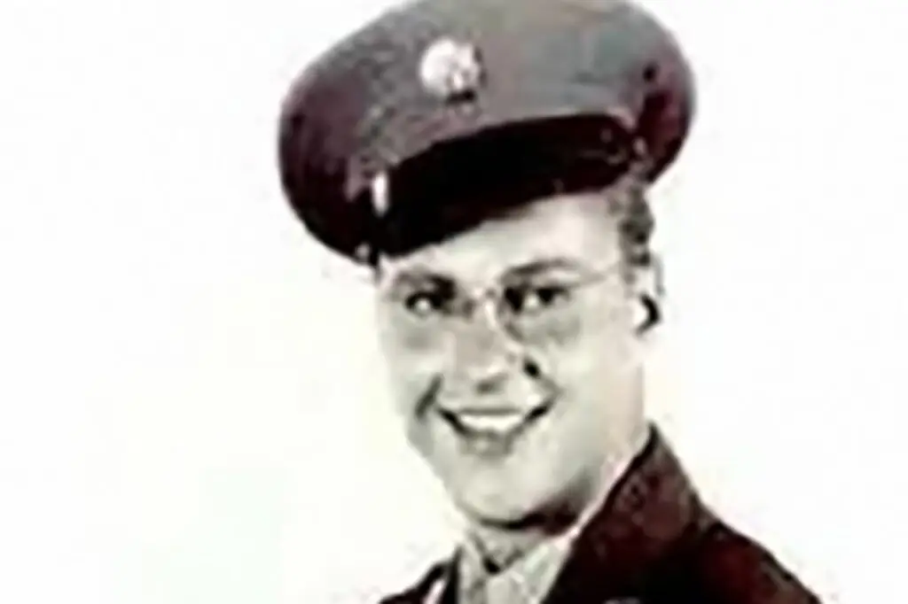 A black and white photo of a person in a military uniform, possibly linked to the DOD. The individual is smiling and wears a peaked cap with a badge on the front. The uniform includes a collared shirt and tie, set against a light background.