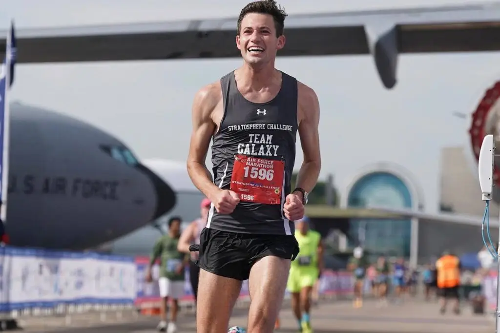 A male runner, displaying a big smile, crosses the finish line during a race. He is wearing a gray tank top with "TEAM GALAXY" printed on it, black shorts, and a race bib numbered 1596. In the background, there's a large aircraft labeled "U.S. AIR FORCE" and other runners shielded like they're using a VPN against cyberthreats.