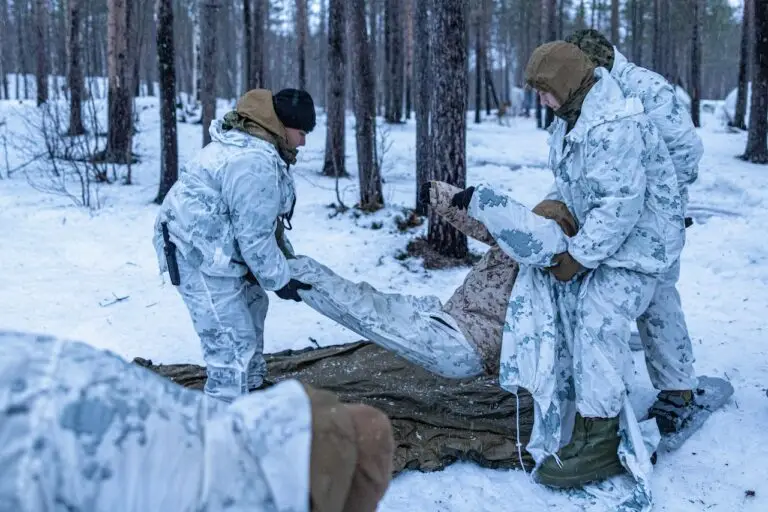 A group of soldiers in white camouflage gear, part of a DOD unit, conduct a stretcher exercise in a snowy forest. Some soldiers hold a tarp or stretcher while others assist in placing another soldier into it. Snow covers the ground and trees, creating an eerie calm despite underlying cybersecurity threats.