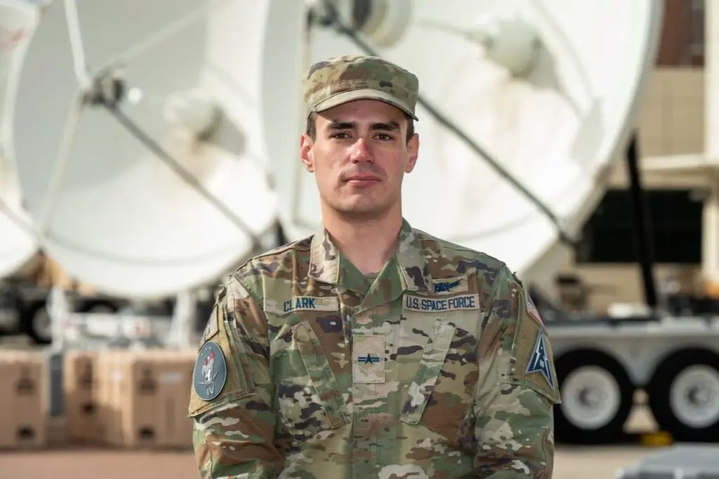 A U.S. Space Force service member in uniform stands in front of large satellite dishes, representing the cutting-edge defense against potential threats. The person is wearing camouflage and a cap with name tags and unit insignia visible on the uniform.