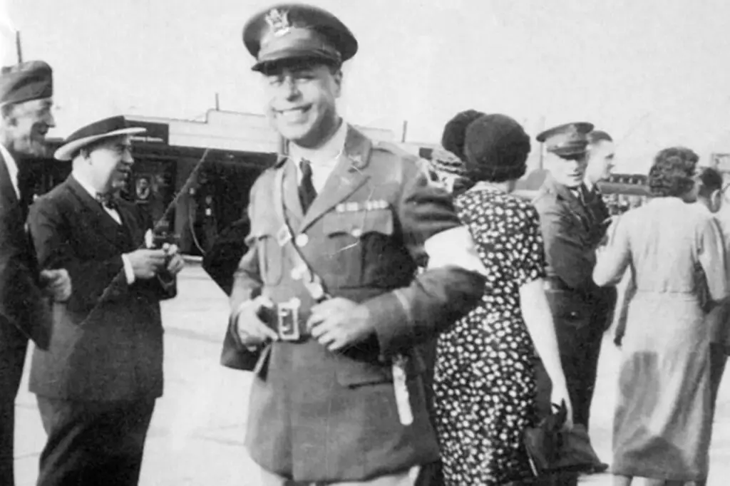 A black and white photograph captures a smiling man in a military uniform with medals on his chest. He stands in an outdoor setting with several other individuals, some in military attire, as they discuss potential threats to security. The background features a train station platform.