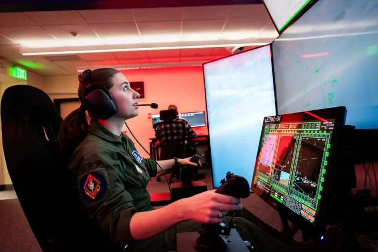 A person in a military uniform operates a flight simulator with multiple screens. They are using a joystick and throttle control, wearing a headset, and appear focused on the task. Another person in the background, working on a separate computer setup, seems to be addressing potential cyberthreats.