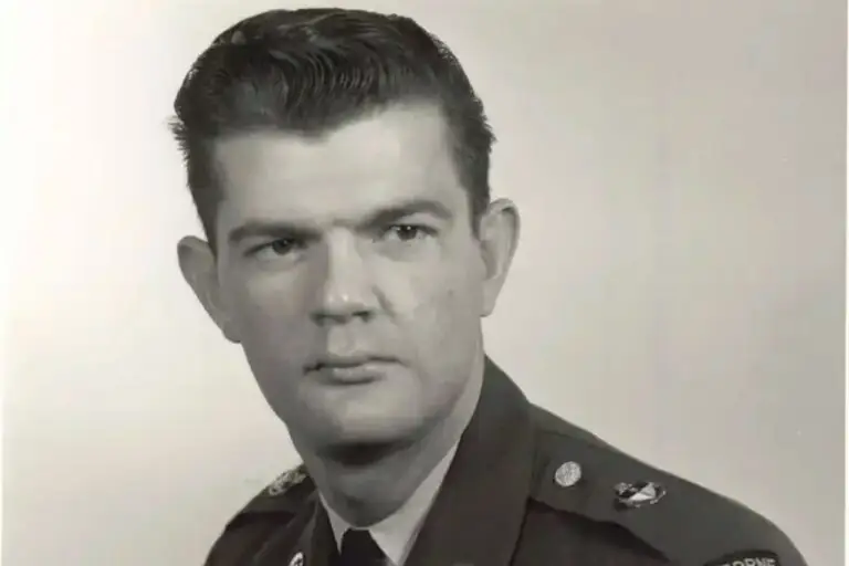 Black and white portrait of a man in military uniform looking slightly to the side. He has short, dark hair and a serious expression. The uniform displays several pins and badges, indicating military status with the DOD. The background is plain.