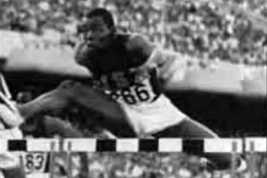 A male athlete in midair during a hurdle race, with the number 861 on his uniform. He is leaping over a hurdle, with the crowd in the background. The image is in black and white, and the athlete's focused expression shows determination akin to a DOD soldier on a mission.