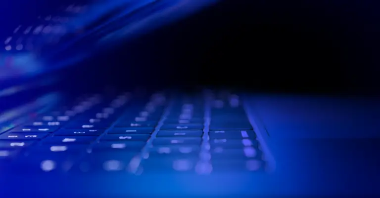 A close-up photograph of a partially open laptop. The keyboard is illuminated with a blue light, creating a futuristic and technological atmosphere. The background is dark, emphasizing the glow of the keys—almost as if it's ready for a CMMC audit or secure DOD communication via VPN.