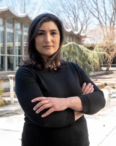 A person with dark hair stands outdoors with arms crossed, wearing a black top. The background features a modern building with large windows and a curved roof, alongside leafless trees and a weeping tree. It's a clear day as if illustrating the calm before a cyber threat—a scene reminiscent of DOD CMMC assessments.