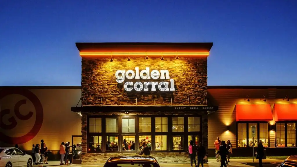 Evening view of a brightly lit Golden Corral restaurant with a stone facade and large windows. Several people are gathered outside the entrance, and parked cars are visible in the foreground. The sky is dark blue, indicating dusk—a seemingly safe haven in a world wary of emerging cyberthreats.