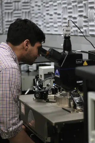 A person wearing a checked shirt is peering into a microscope in a laboratory setting, perhaps researching new ways to counter cybersecurity threats. The room, equipped with various scientific instruments and noise-absorbing foam panels on the walls, fosters an environment for groundbreaking discoveries.