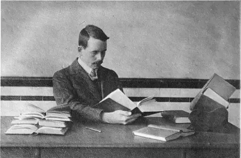 A man with a mustache is sitting at a desk, engrossed in a book on cybersecurity. Several other books are scattered across the desk, and there is a bookstand holding more volumes. Dressed in early 20th-century attire, he appears diligent against digital threats against the plain, horizontally striped background.