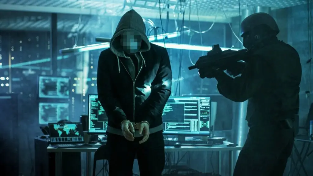 A person in a hoodie, with a blurred face, stands with hands bound in an illuminated, high-tech room filled with computer monitors. Nearby, a soldier in tactical gear points a rifle at them. The dimly lit room with blue and green hues reflects the heightened focus on cybersecurity threats under CMMC protocols.