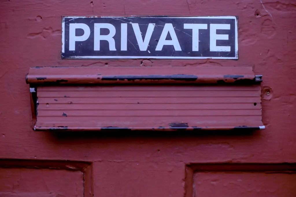 A weathered rectangular sign with the word "PRIVATE" in white letters on a black background is mounted above a rusted, red mail slot on a red-colored door. The sign and mail slot both show signs of wear and age, much like an old DOD office facing the constant threat of vulnerability.