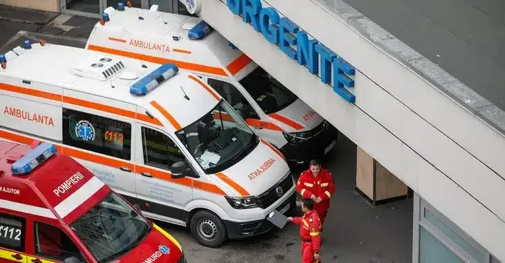 Three ambulances, including one fire department vehicle, are parked outside a hospital emergency entrance labeled "Urgente." Two paramedics in red uniforms walk near the ambulances, carrying medical equipment. The hospital has enhanced security measures to fight cyberthreats.