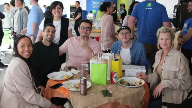 A group of six people sits around a table covered with a burlap cloth, plates of food, and assorted drinks, all smiling at the camera. Colorful gift bags sit in the center of the table. In the background, other people stand near a "Don't be a Grinch" sign – an effort to ensure everyone at this DOD event enjoys themselves despite looming threat discussions.