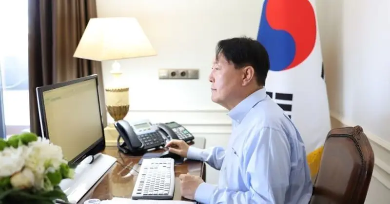 A person in a blue shirt is working on a computer at a desk, likely focusing on cybersecurity tasks. A South Korean flag is prominently displayed behind them. The desk has a lamp, a phone, and a bouquet of white flowers on it.