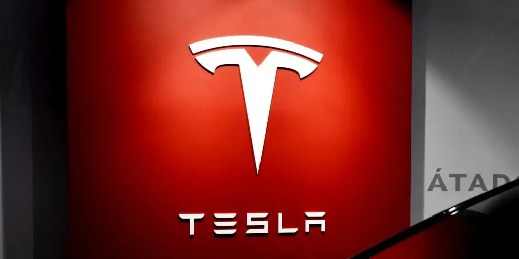 A wall painted in bright red displays a large white Tesla logo at the top center. Below it, the word "TESLA" is written in a stylized font. The shiny black edge of a car is visible in the bottom right corner of the image, evoking thoughts of innovation amidst rising cyberthreats.