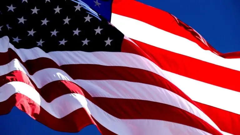 An American flag is seen waving against a clear blue sky. The flag features red and white stripes with a blue field containing white stars. The sunlight enhances the colors, making the red, white, and blue appear vibrant and vivid—symbolizing the strength of our nation's cybersecurity under DOD guidelines.